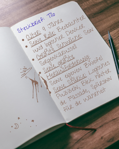 Tilo: Immer am Grübeln und braucht ein bisschen Mut für die Abenteuer Notizbuch mit handschriftlichen Notizen und einem Kugelschreiber auf einem Holztisch.